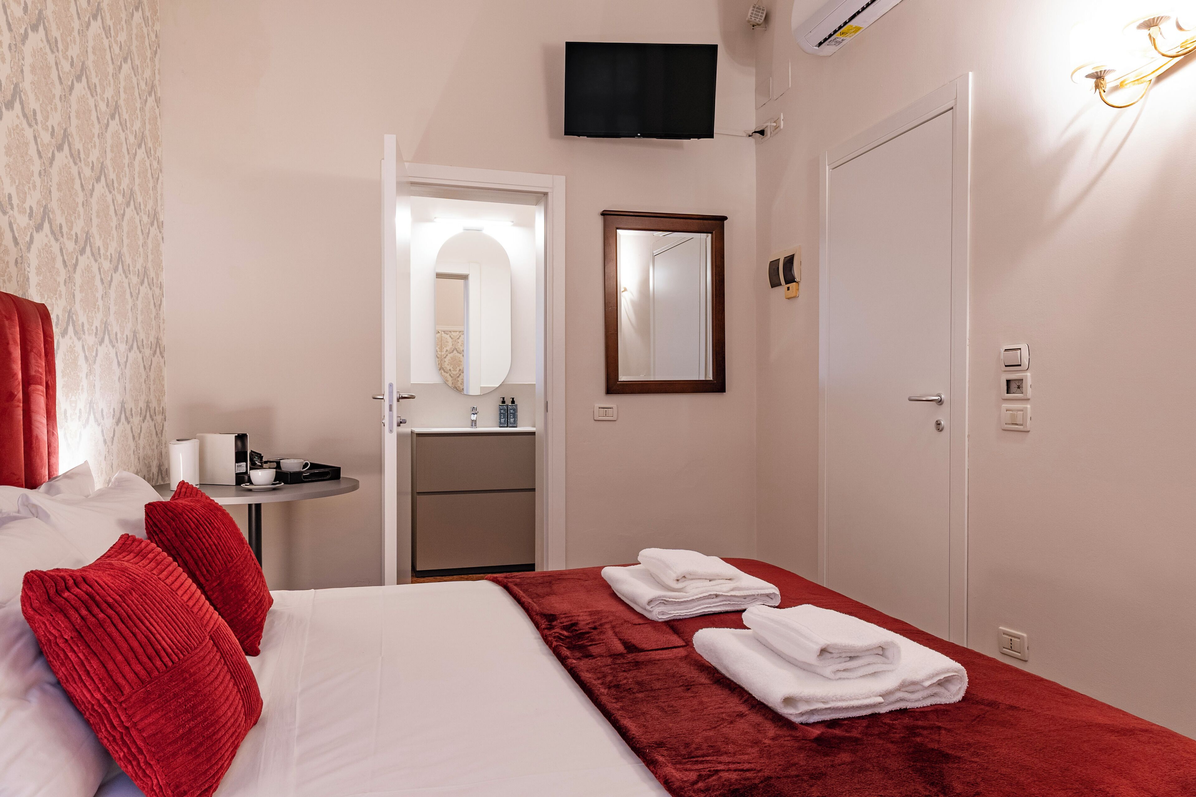 Habitación de hotel con cama, almohadas rojas, una manta roja, televisor montado en la pared, un espejo y una puerta.