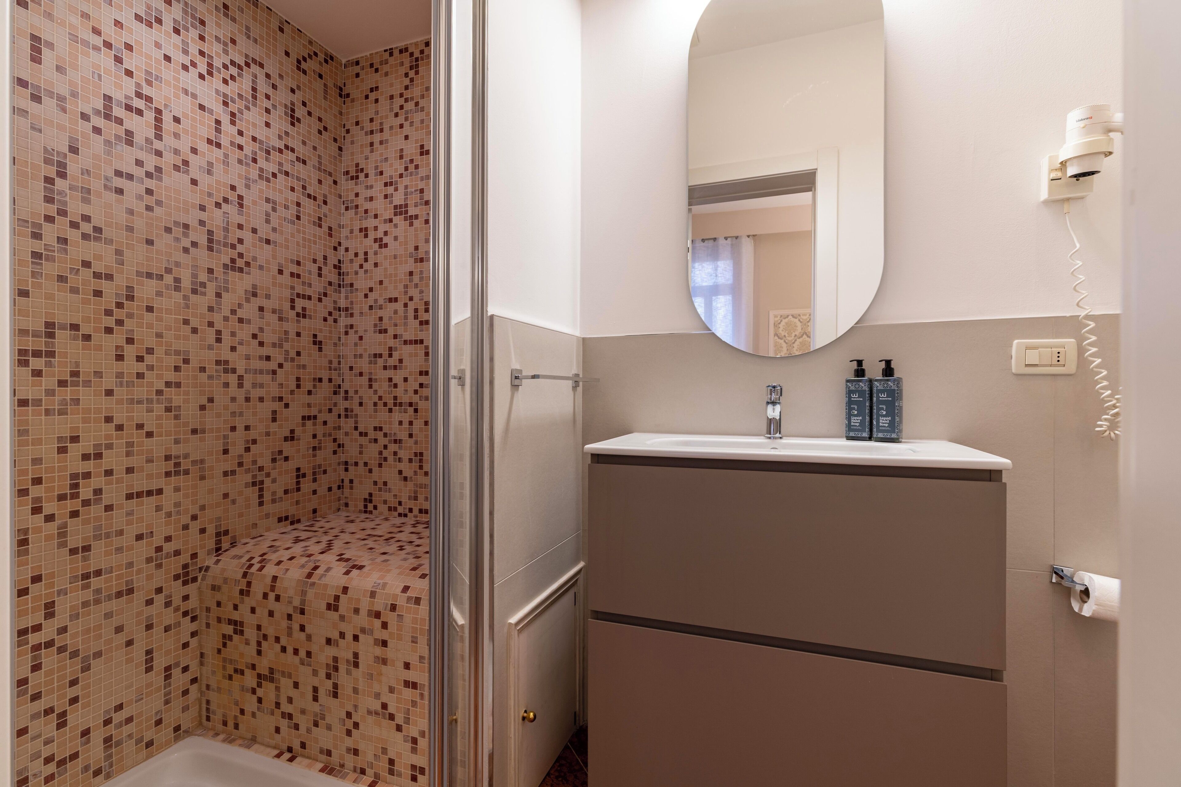 A bathroom with a tiled shower, a sink with two bottles, and a mirror.