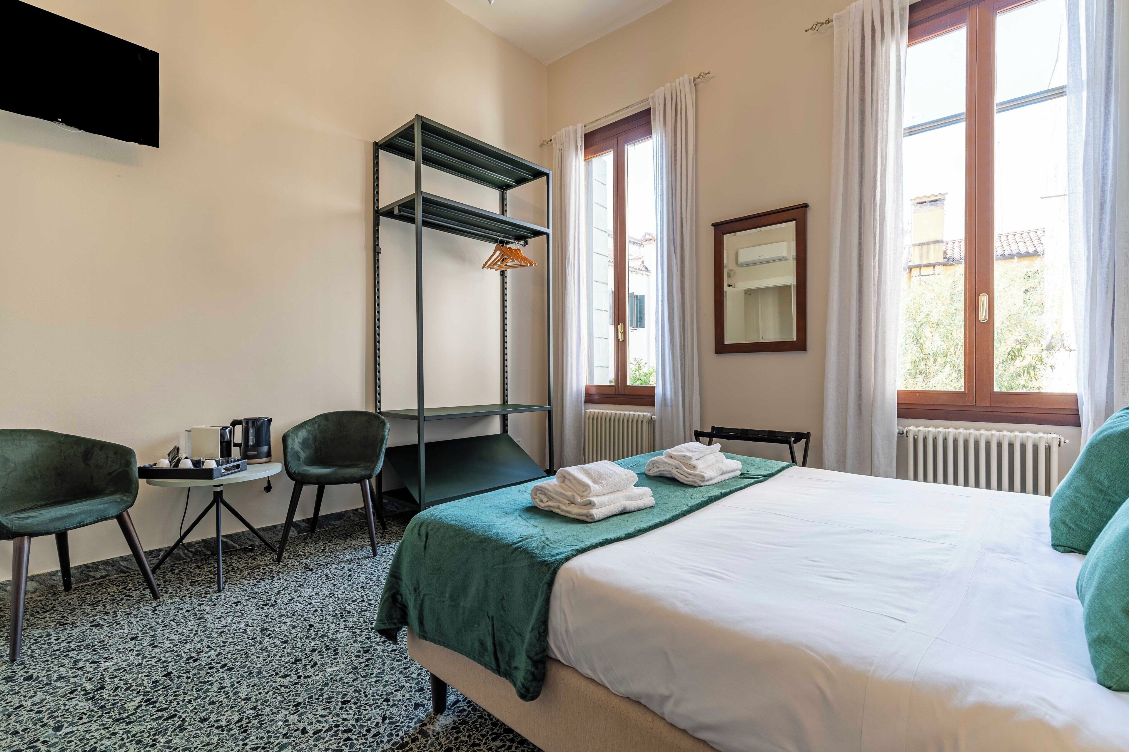 A hotel room with a bed, a desk, two chairs, a small table with a kettle, and a radiator.