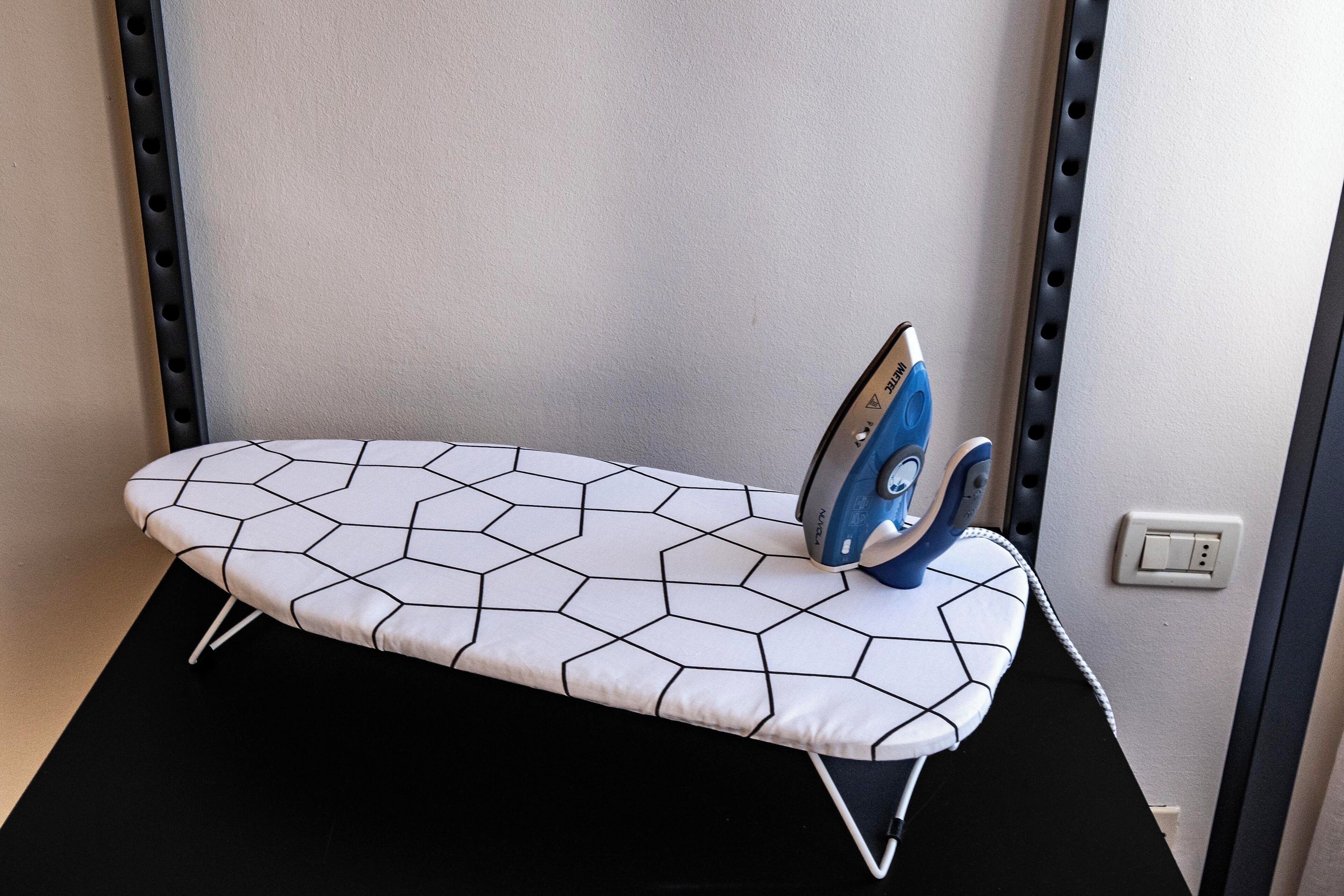 Ein kompakter Bügelplatz mit einem Bügelbrett mit geometrischem Muster, einem blauen Bügeleisen und einer Steckdose an der Wand.