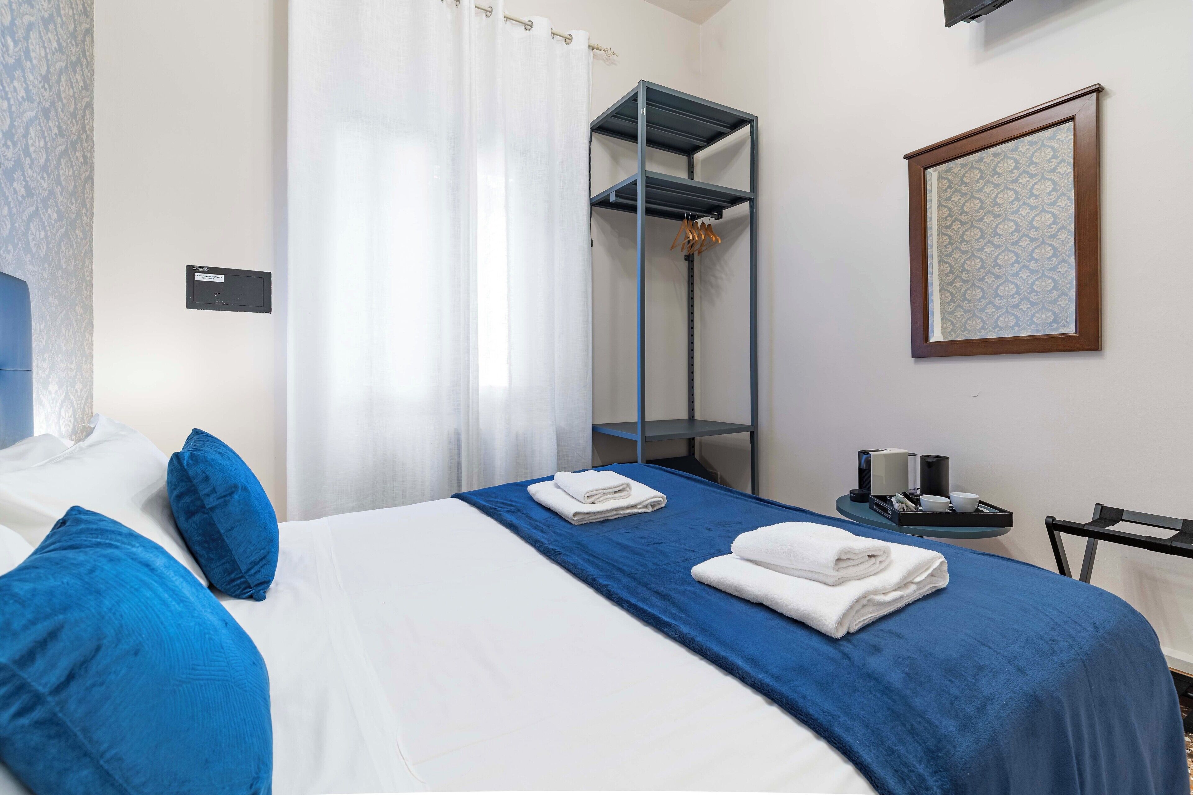 Quarto de hotel com cama, colcha azul, travesseiro branco, travesseiro azul, manta azul, toalha branca, espelho, moldura de quadro, mesa pequena com bandeja e cadeira.