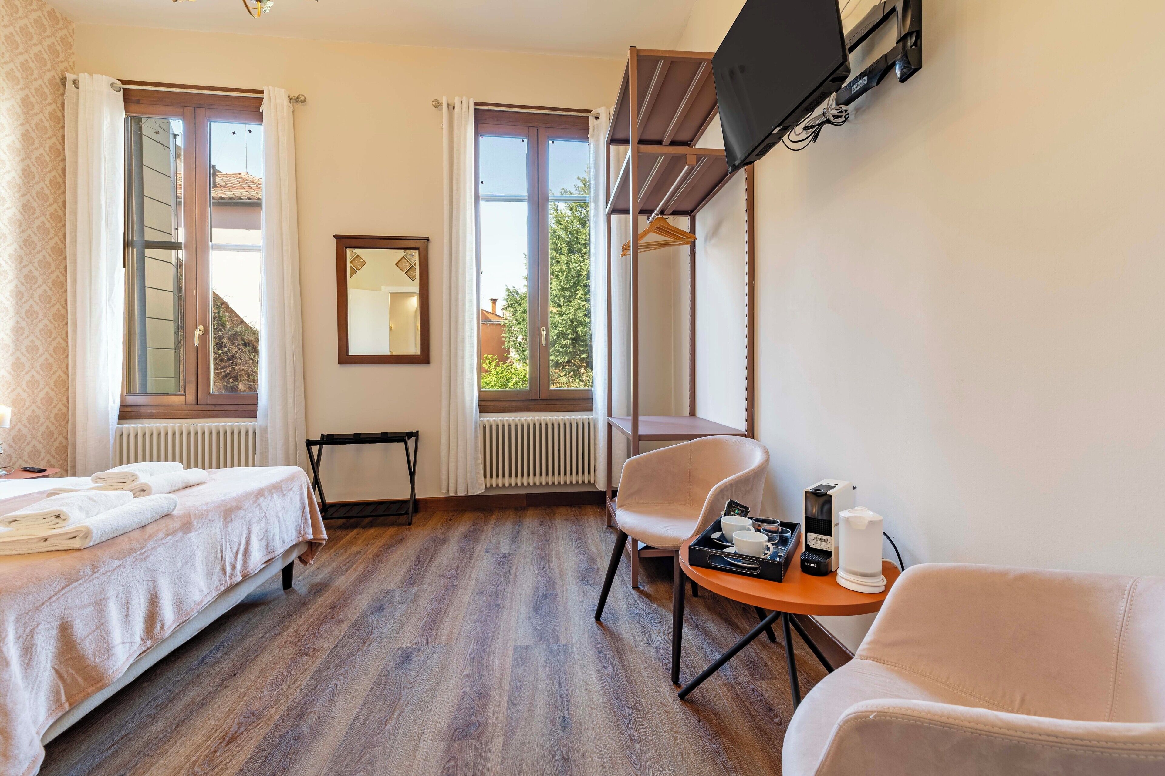 Habitación de hotel con cama, dos sillones, una mesita con detalles de bienvenida, televisor montado en la pared y ventanas amplias.