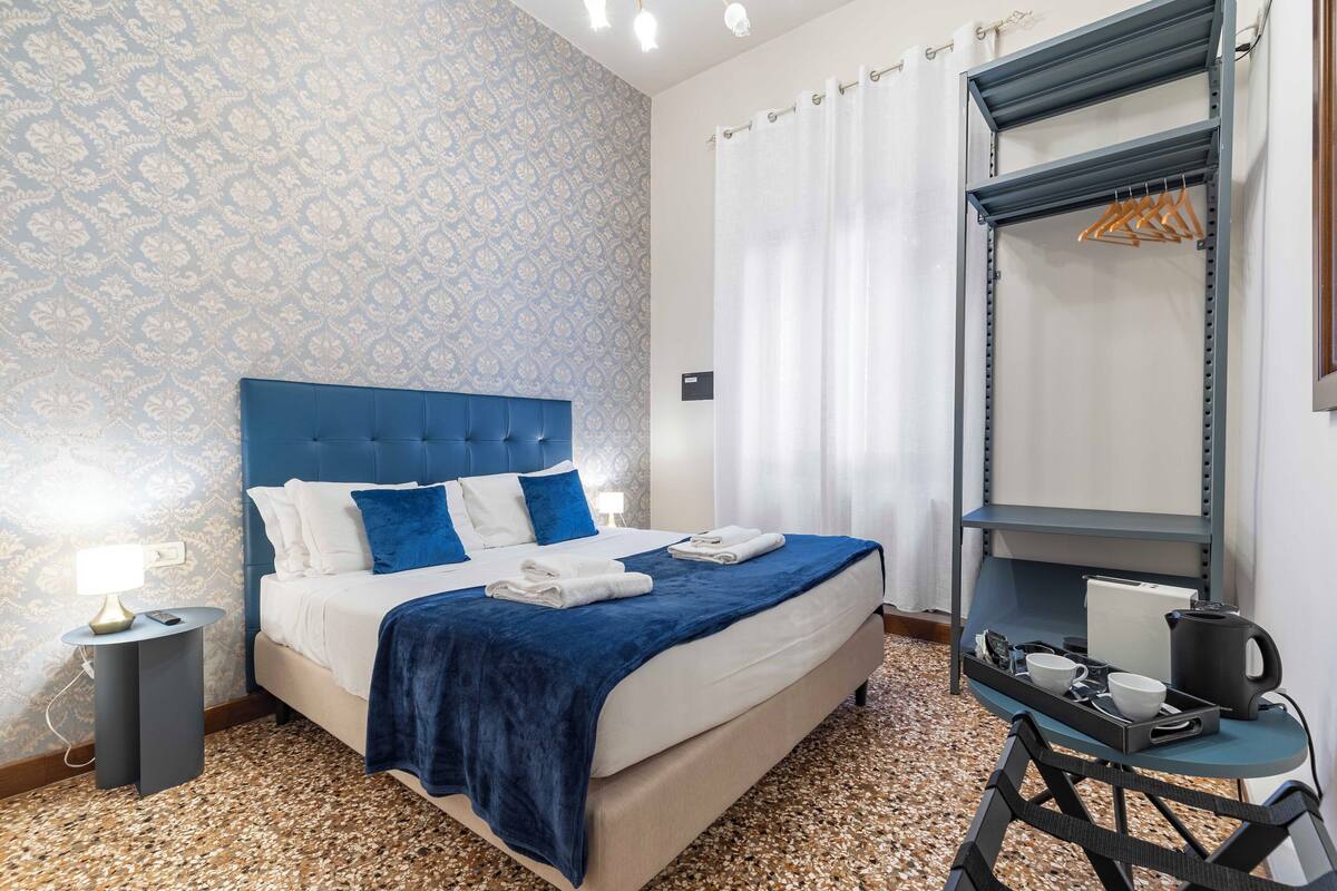 Ein Hotelzimmer mit einem Bett, einem Nachttisch mit Lampe, einem kleinen Tisch mit Tablett und einem Schrank mit Kleiderhaken.