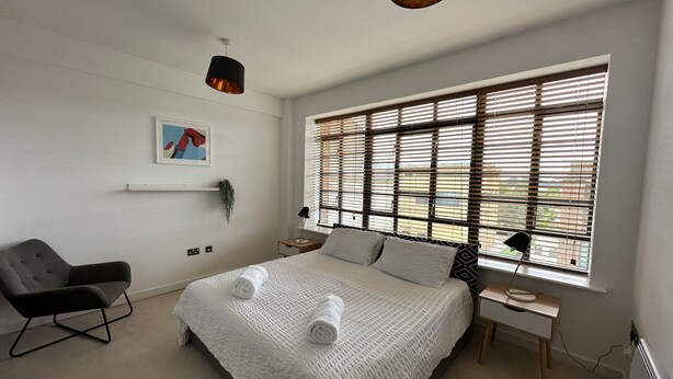 A modern bedroom with a large bed, a chair, a bedside table, and a window with blinds. - Travaal.com - FREE-WIFI-NETFLIX-PARKING - 2 Bedroom Apartment - Farnborough (Farnborough)