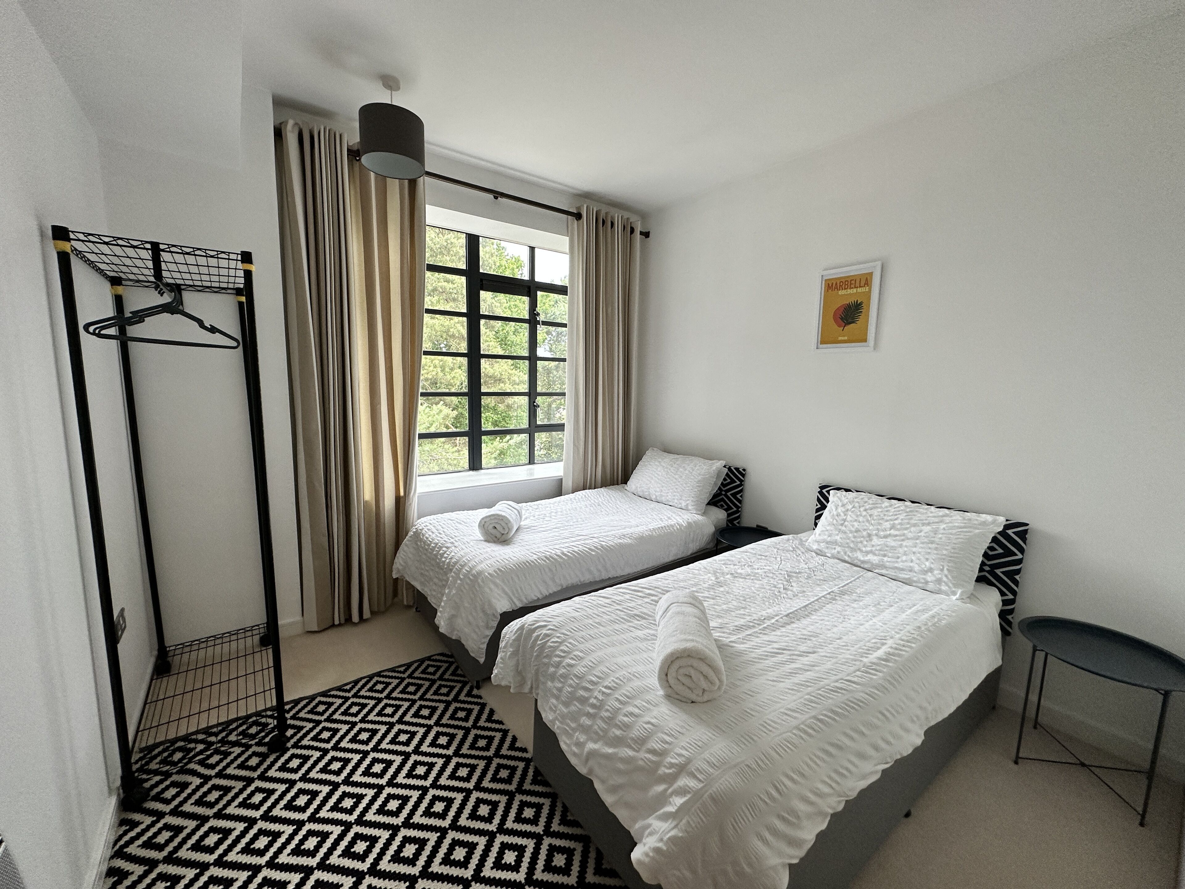 Ein Hotelzimmer mit zwei Betten, einem Fenster mit Vorhängen, einer Pendelleuchte, einem gemusterten Teppich und einem gerahmten Bild an der Wand.