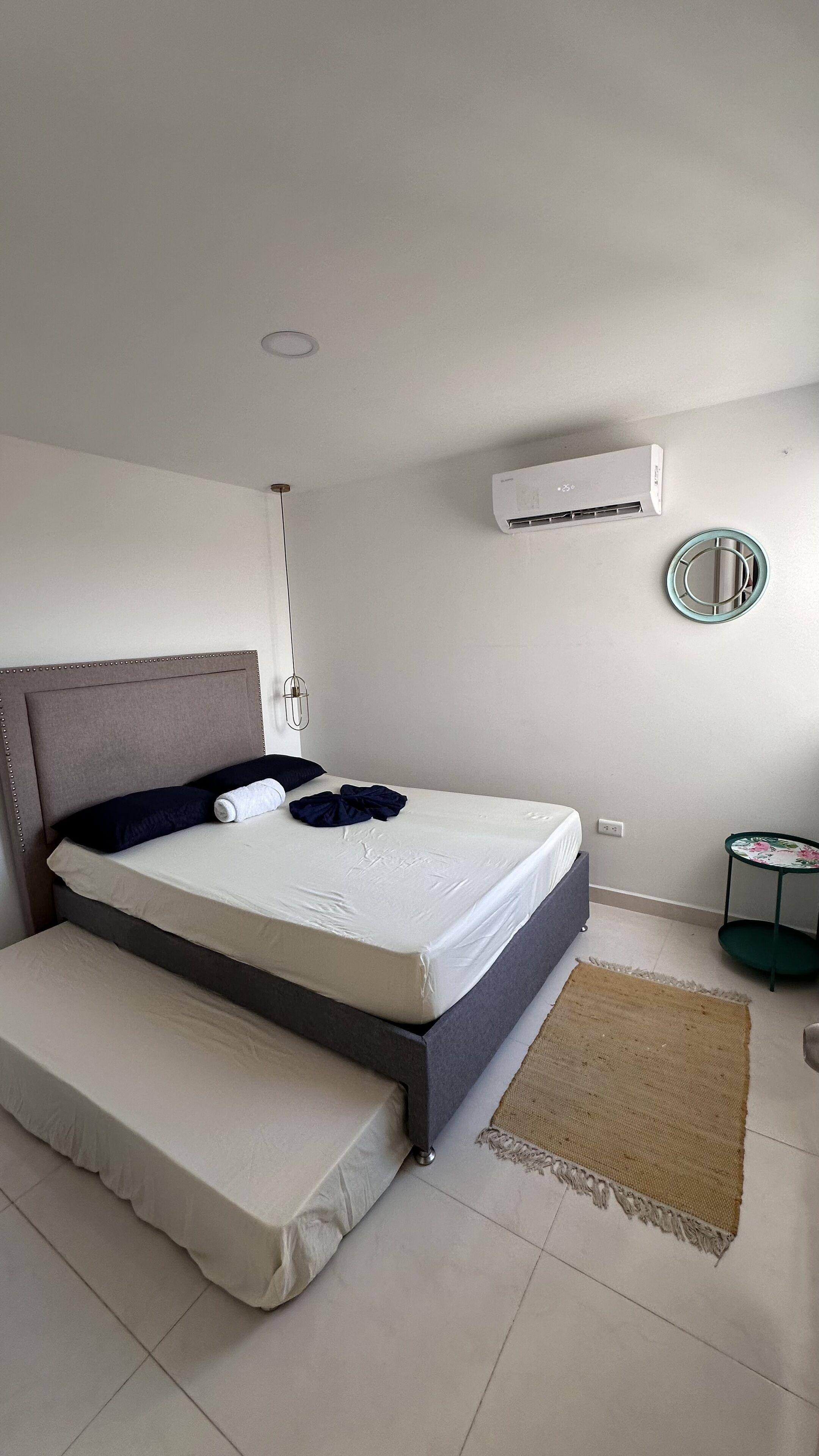 Een netjes opgemaakt bed met hoofdbord, een klein nachtkastje, een wandairconditioner en een ronde spiegel.