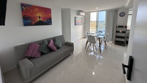 A modern living room with a grey sofa, colorful cushions, a dining table with chairs, and a large window offering a view of the outdoors.