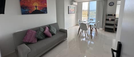 Een moderne woonkamer met een grijze bank, kleurrijke kussens, een eettafel met stoelen en een groot raam met uitzicht op het landschap.