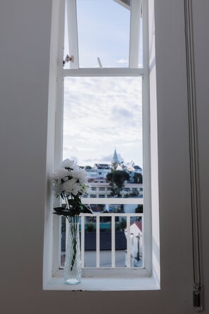 Une fenêtre avec un vase blanc contenant des fleurs blanches, et une vue sur des immeubles et le clocher d’une église à l’extérieur.