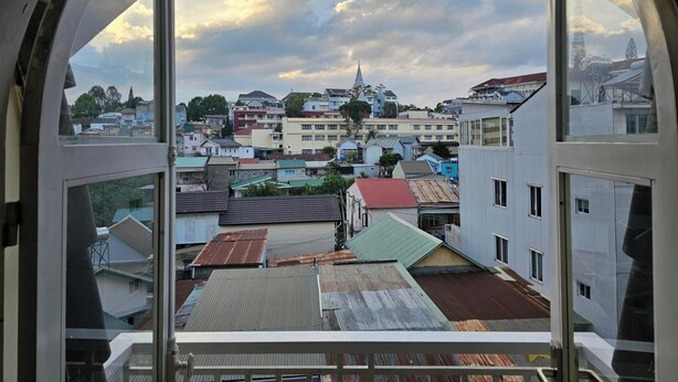 A view through a window with a white frame, overlooking a cityscape with various buildings, including a church with a steeple, under a partly cloudy sky. - 42 Da Lat Hotel (Xuan Huong - Da Lat)