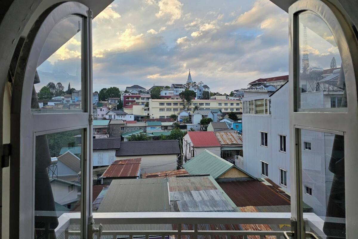 A view through a window with a white frame, overlooking a cityscape with various buildings, including a church with a steeple, under a partly cloudy sky.