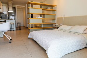 A modern hotel room with a large bed, a desk with a chair, and a kitchenette in the background.
