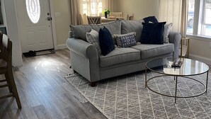 Een woonkamer met een grijze bank, een tapijtend met een patroon en een glazen salontafel.