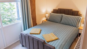 A bedroom with a bed, two pillows, a nightstand with a lamp, a window with curtains, and a radiator. - 2 Bed Barn conversion (Tibenham)