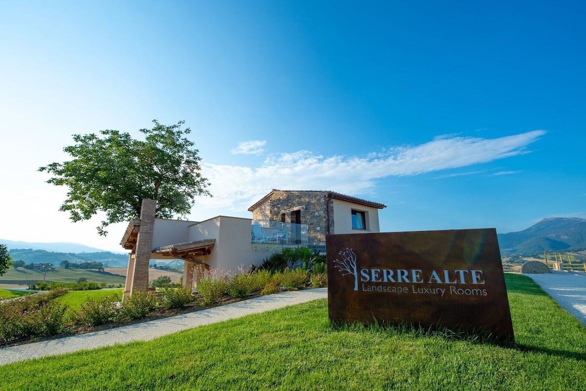 Une enseigne indiquant « Serrre Alte Landscape Luxury Rooms », avec en arrière-plan une vue sur un bâtiment moderne et, au premier plan, un arbre.