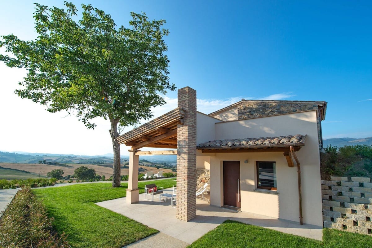 Une maison moderne avec une cheminée en pierre, un patio couvert et une vue sur des collines ondoyantes.
