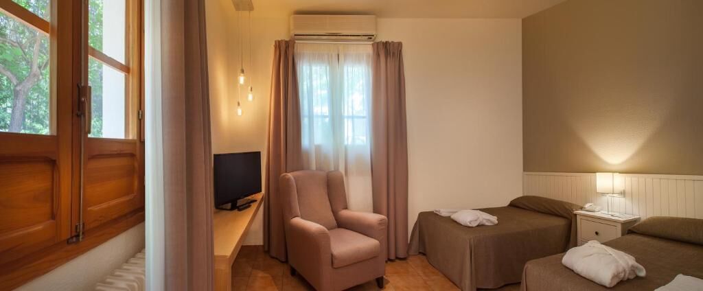 Ein Hotelzimmer mit Holzboden, einem Einzelbett, einer Schlafcouch, einem Sessel, einem Fernseher und einem Fenster mit Vorhängen.