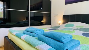 A modern bedroom with a large window, a bed with blue and green bedding, a nightstand with a lamp, and a painting on the wall.
