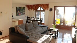 A living room with a sofa, a dining table with chairs, a kitchen area, and a large window with a view.