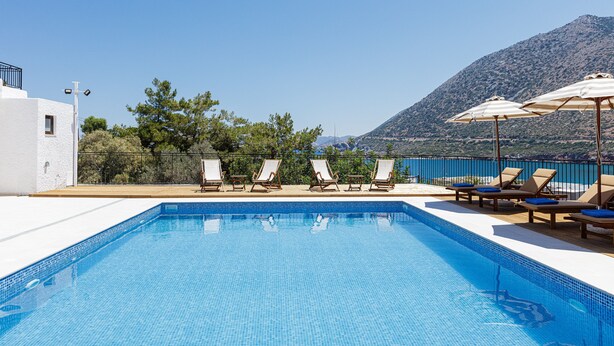 Una piscina con lettini e ombrelloni, circondata da un edificio bianco e una montagna sullo sfondo.