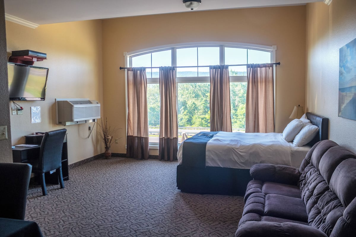 Ett hotellrum med en säng, en soffa, ett skrivbord med en stol, en tv och ett fönster med gardiner.