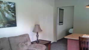 Un salon avec un canapé, une lampe, une petite table et une chaise. Il y a un tableau au mur et un miroir sur la porte.