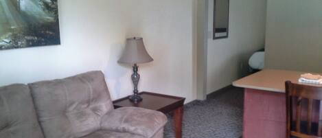 A living room with a sofa, a lamp, a small table, and a chair. There is a painting on the wall and a mirror on the door.
