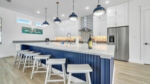 Una cocina moderna con una isla central, taburetes de bar, iluminación colgante y un refrigerador de acero inoxidable.