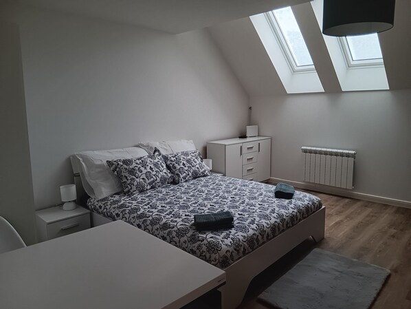 A bedroom with a bed, a desk, a nightstand, a radiator, and two skylights.