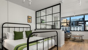 A modern bedroom with a large window, a bed with white bedding and green pillows, a small round table, and a view of the cityscape. - Stylish Minimalist: The Tokyo|Mountain and City View (Cape Town)