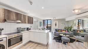 A modern kitchen with a central island, built-in oven, and microwave. There is a dining area with a round table and chairs. The living room features a gray sofa and ottomans. - Modern Apartment in Trendy De Waterkant (Cape Town)