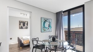 A modern dining area with a glass table and black chairs, a cityscape painting, and a view of a building through large windows. - Modern Apartment in Trendy De Waterkant (Cape Town)