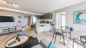 A modern living room with a sofa, coffee table, and TV. There is a kitchen area with a fridge, stove, and bar stools. - Modern Apartment in Trendy De Waterkant (Cape Town)