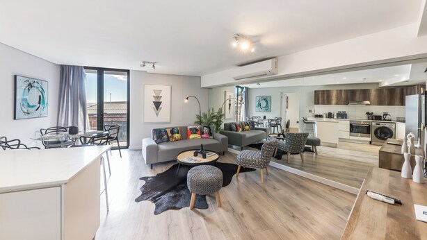 A modern living room with a sofa, coffee table, and ottoman, a dining area with chairs, and a kitchen with appliances. - Modern Apartment in Trendy De Waterkant (Cape Town)