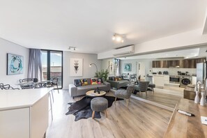 Una sala moderna con sofá, mesa de centro y reposapiés, un comedor con sillas, y una cocina equipada.