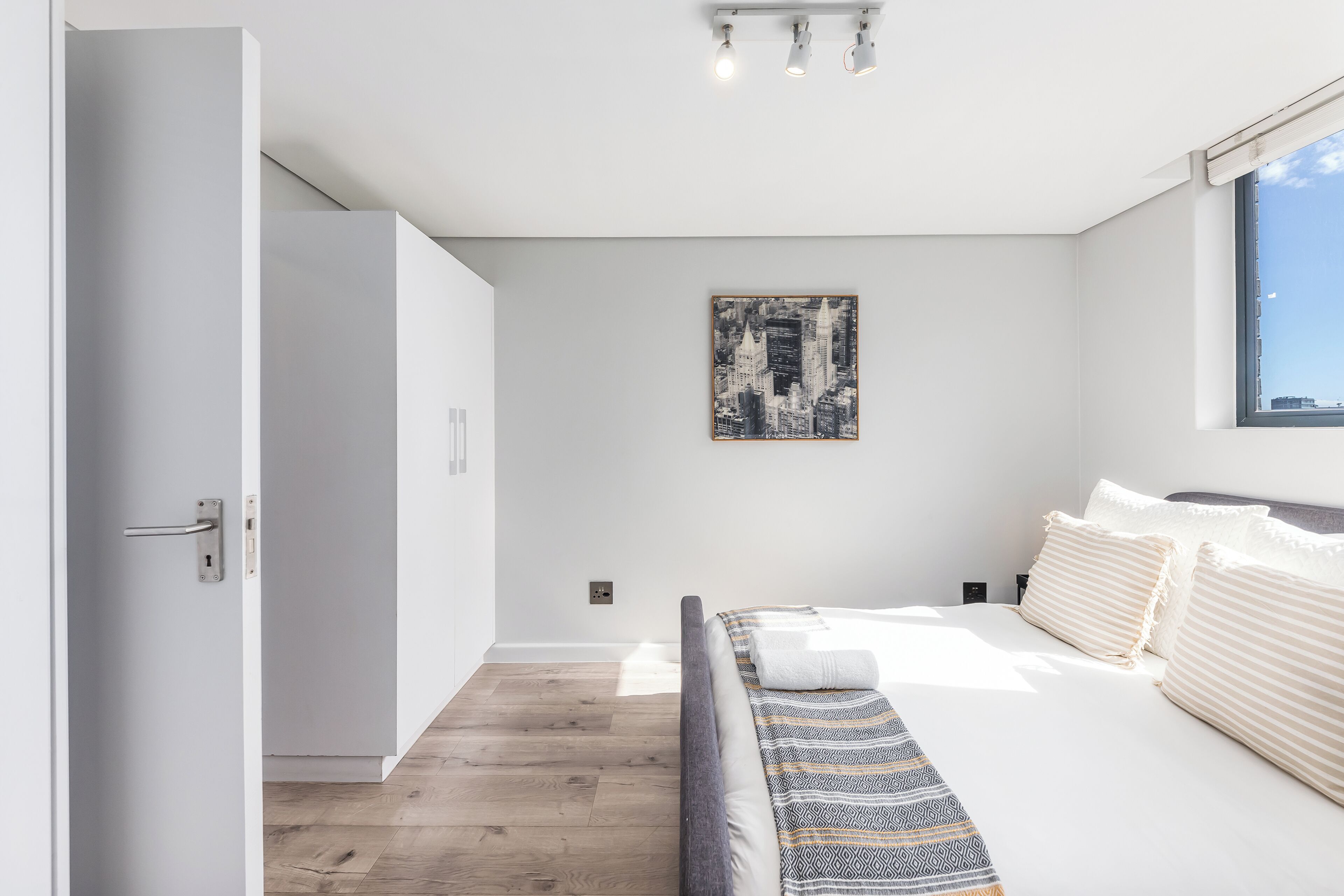 A modern bedroom with a large bed, a cityscape painting on the wall, and a view of the sky through a window.