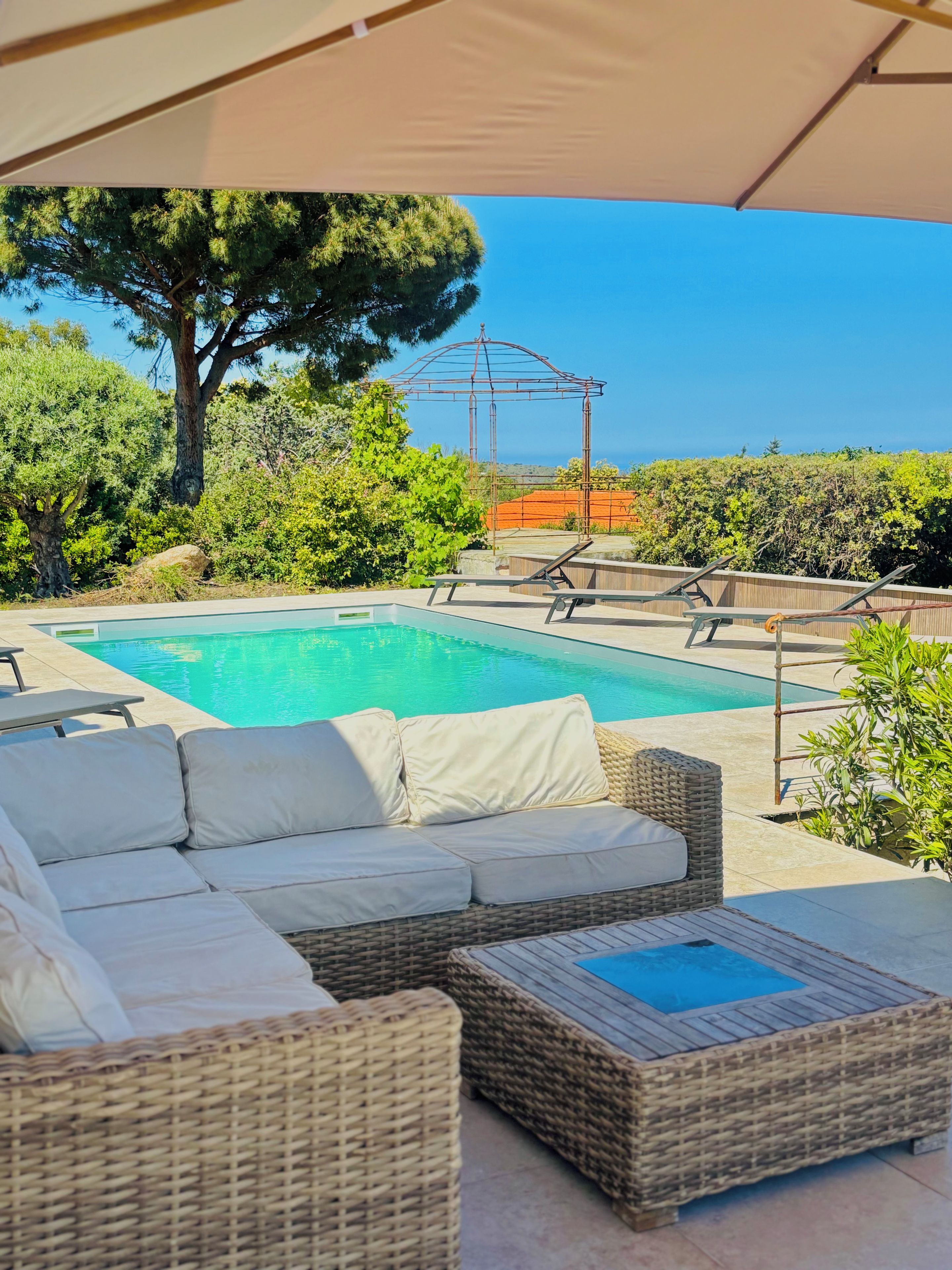 Zona piscina con divano in vimini, una poltrona e una struttura a pergola.