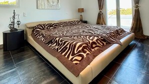 A spacious bedroom with a large bed, a nightstand with a lamp, a wall-mounted artwork, and a view through the window. - Villa de 220 m2 Avec Piscine et vue mer à 5min des Plus Belles Plages de Balagne (L'ile-Rousse)