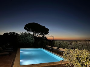 A swimming pool with lounge chairs and a pergola, surrounded by trees and a view of the horizon.