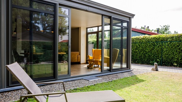 Ein modernes Haus mit großen Glasüren, die auf eine Terrasse mit Liegestuhl und gelbem Ohrensessel führen.