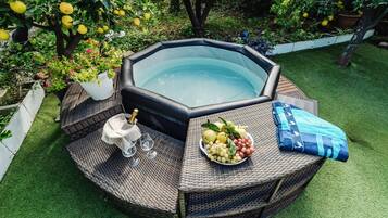 Un bain à remous entouré d’une table en osier, sur laquelle se trouvent un panier de fruits et une bouteille de champagne.