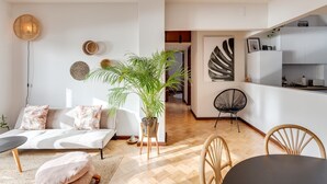 Een moderne woonkamer met een bank, een eettafel met stoelen, een grote plant in pot en een keuken met kastjes en een radio.