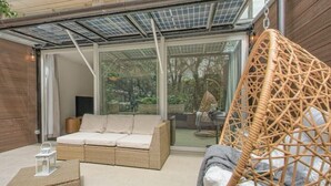 A modern outdoor living space with a wicker sofa, a hanging chair, and a glass door leading to a garden. - King-size bed with terrace and Air Conditioning!
 (Las Condes)