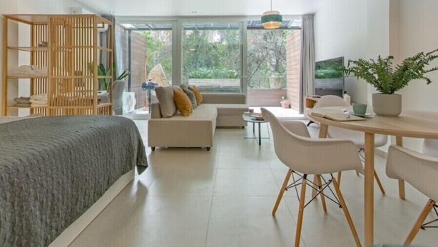 A modern, well-lit room with a bed, sofa, dining table, and chairs. There is a large window providing a view of greenery outside. - King-size bed with terrace and Air Conditioning!
 (Las Condes)