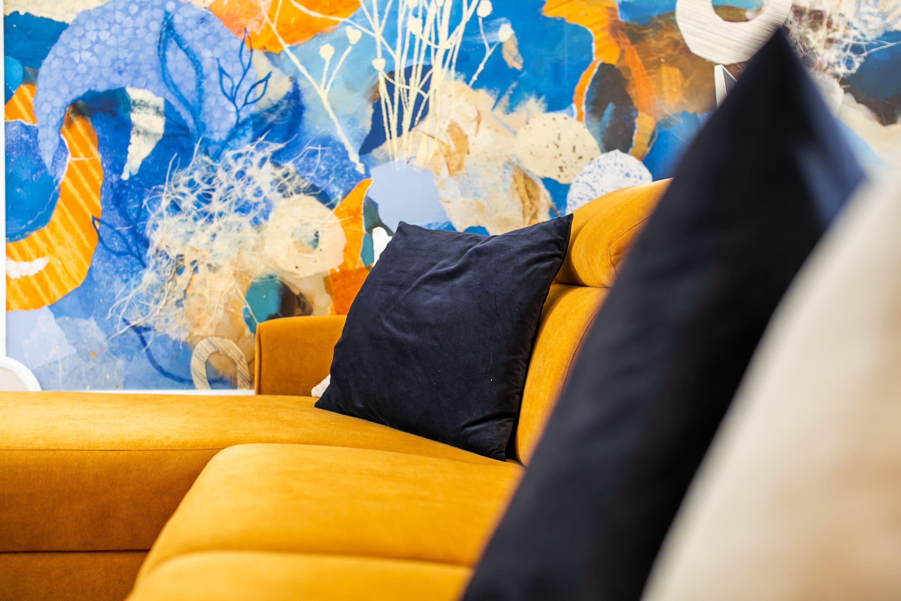 A modern living room with a yellow sofa, a dark blue pillow, and a colorful abstract painting on the wall.