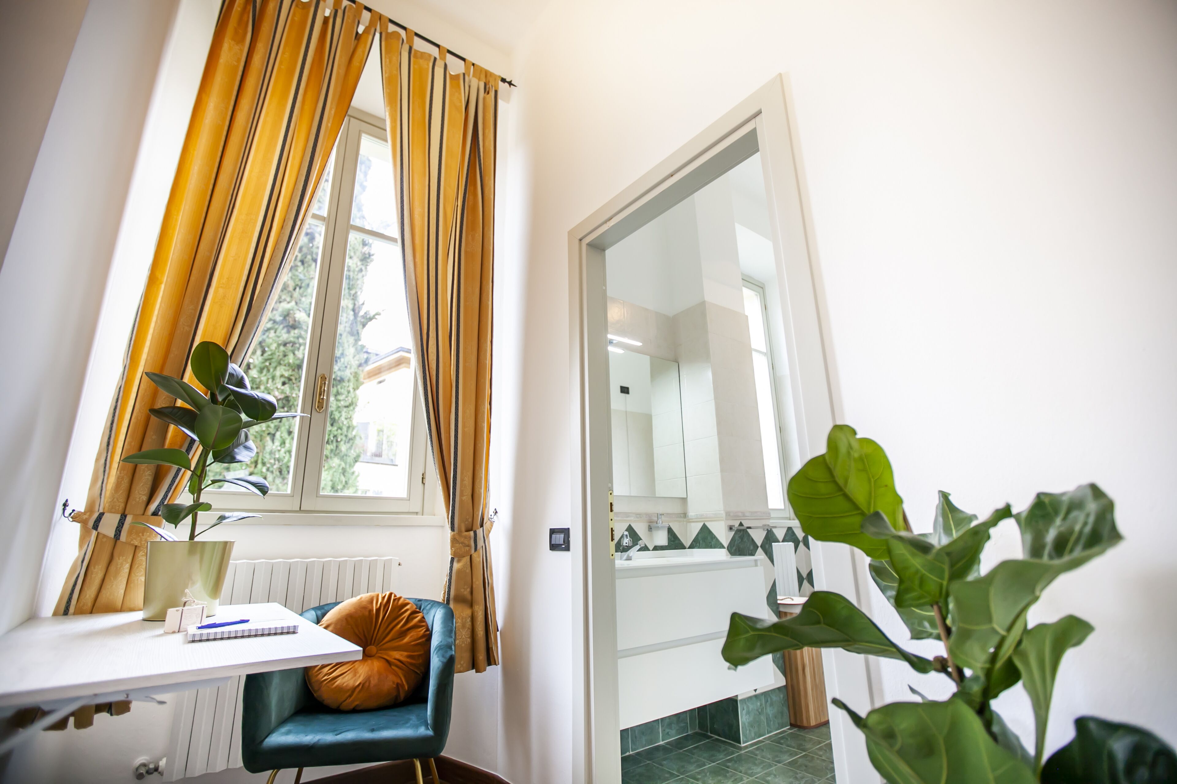 A room with a window, a desk with a chair, a plant, and a door leading to a bathroom.