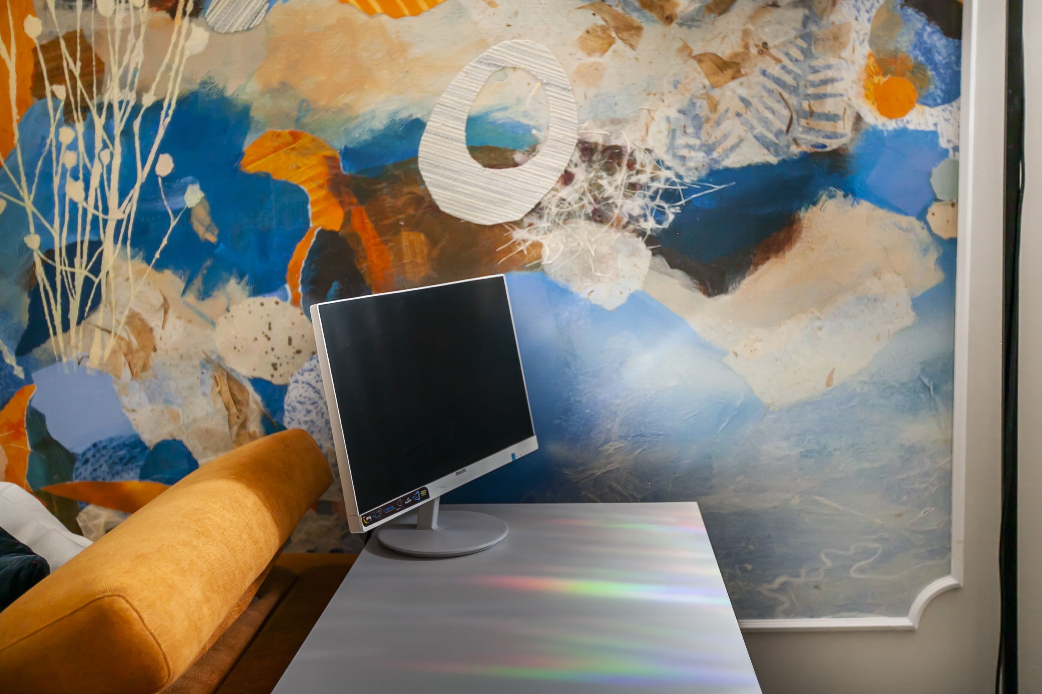 Een moderne kamer met een groot abstract schilderij aan de muur, een flatscreen monitor op een witte tafel en een houten stoel.