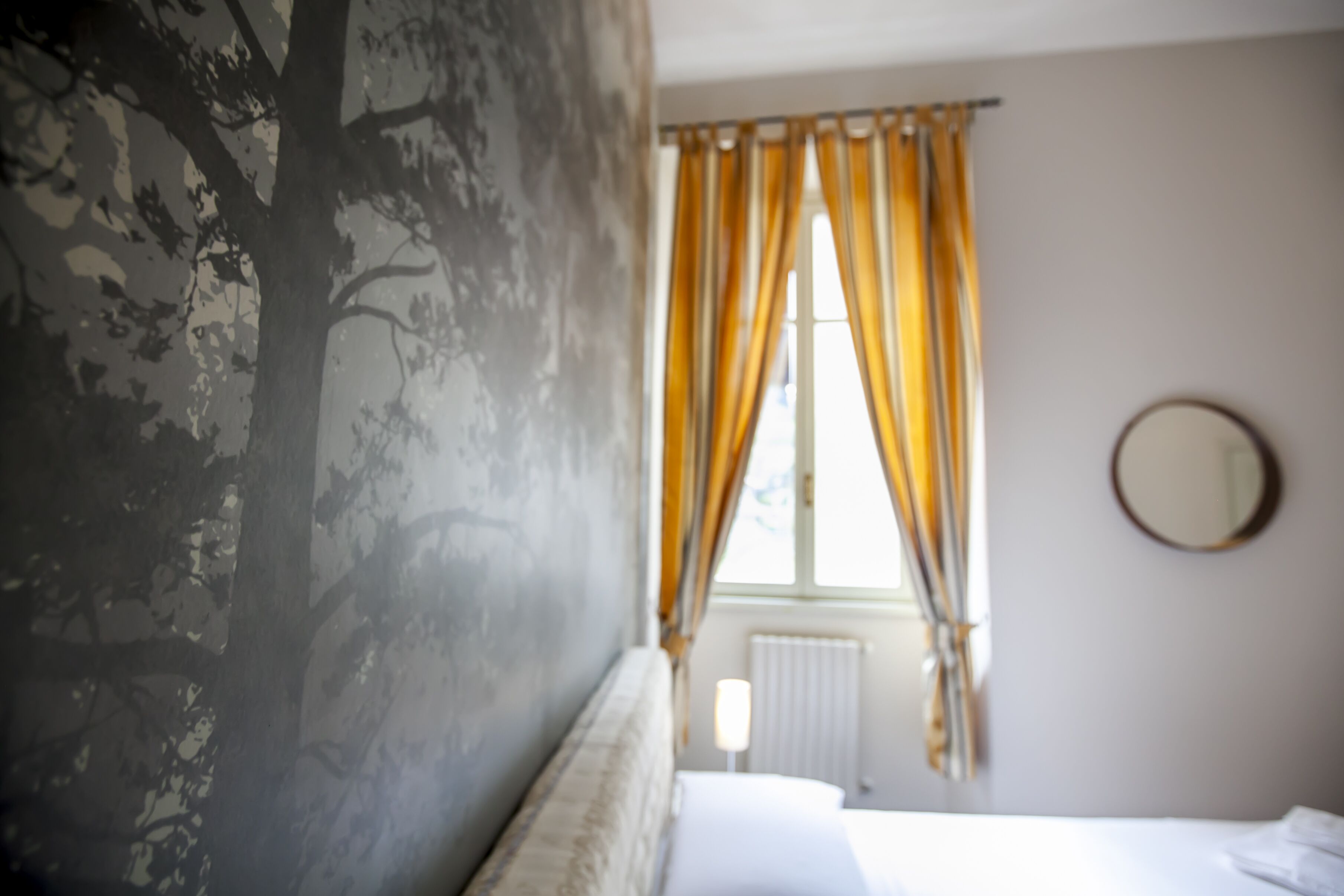 A bedroom with a bed, a window with curtains, a radiator, and a round mirror on the wall.