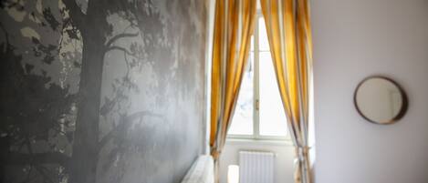 A bedroom with a bed, a window with curtains, a radiator, and a round mirror on the wall.