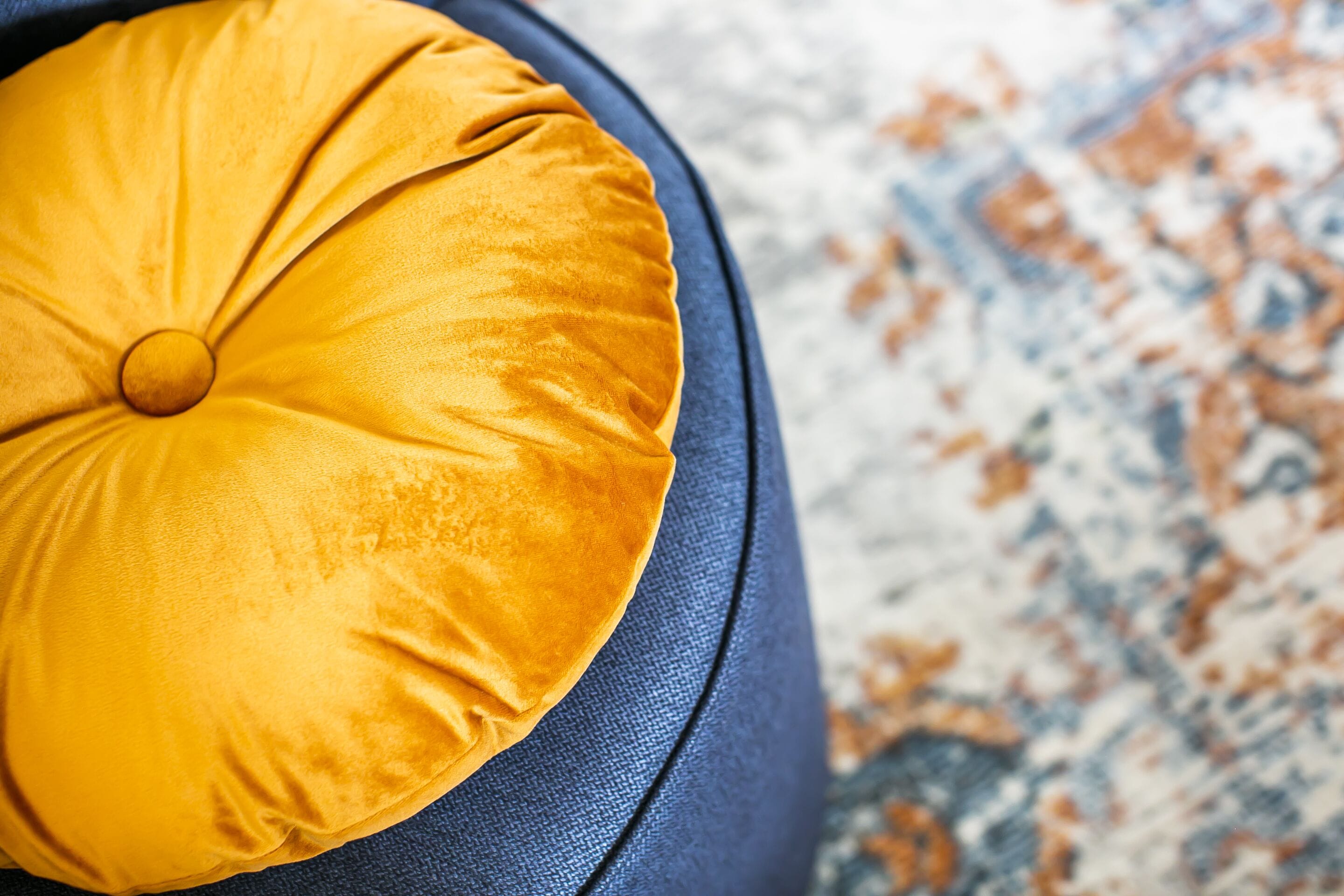 A close-up of a yellow cushion on a dark blue sofa with a patterned rug in the background.